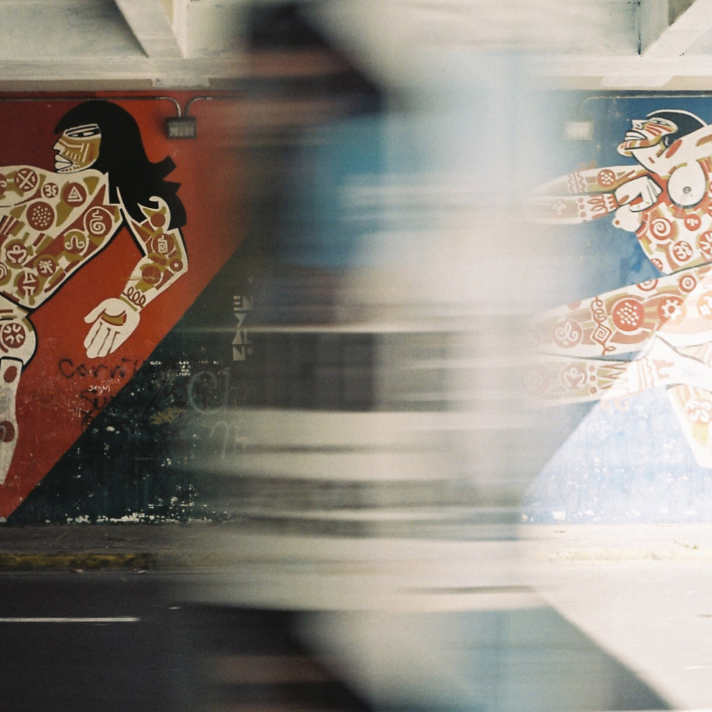 A mural of two stylized figures in dynamic poses, echoing the Weathered brand, is painted under a bridge and partially hidden by a blurred passerby in the foreground.