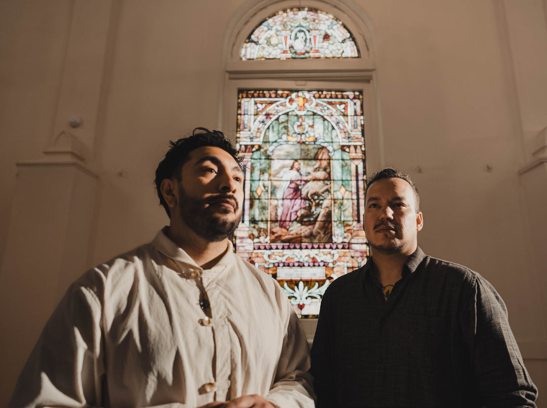Two men stand in front of a tall, colorful stained glass window inside a church. They appear thoughtful, with soft light illuminating their faces and the intricate window behind them.